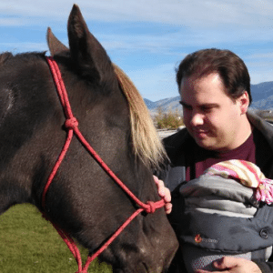 Matt Gowdy pets a brown horse while holding a baby in a baby carrier